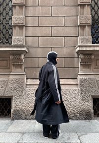 Personne portant un long manteau noir et un bonnet tricoté marchant devant un bâtiment en pierre texturée avec des fenêtres décoratives sur le trottoir de la ville.