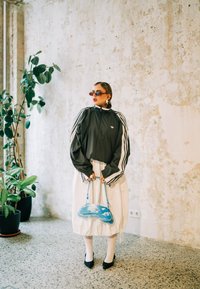 Casaco desportivo cinzento com riscas brancas, saia branca, mala azul com design de nuvem, sapatos de salto pretos e óculos de sol, contra uma parede texturizada.