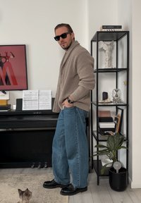 Cardigan beige côtelé, jean bleu oversize et chaussures noires. Accessoires comprenant des lunettes de soleil et une montre. En arrière-plan, une étagère à livres et un piano.