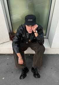 Zwarte leren jas, zwarte Carhartt-cap, bruine corduroy broek, glanzende zwarte schoenen. Zittend tegen een glazen raam. Grijsachtige stoep.