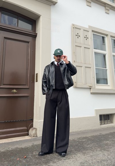 Hombre con chaqueta de cuero negra, pantalones anchos negros, suéter negro, camisa blanca, corbata roja y gorra verde, de pie junto a un edificio blanco con puerta y contraventanas marrones.