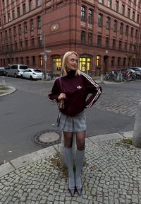 Vinrød lynlåstrøje med hvide striber, grå ternet nederdel, grå knæstrømper og bordeaux spidse hæle, stående på brostensbelagt gade.