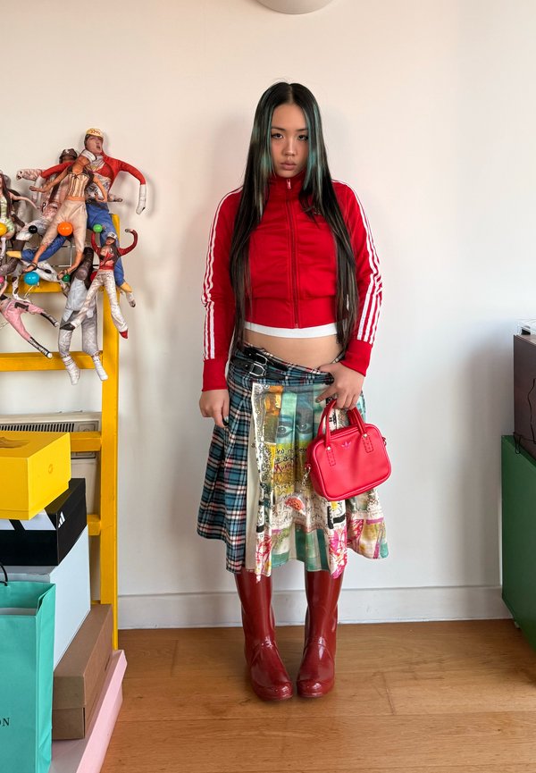 Jeune femme portant une veste courte rouge, un kilt à motifs, des bottes rouges, tenant un petit sac à main rouge, debout à l'intérieur près d'une étagère jaune avec des poupées.