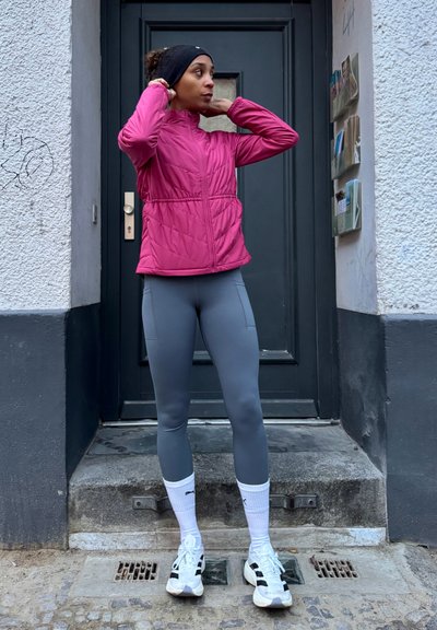 Chaqueta rosa ajustada e aislada con cuello alto, combinada con mallas gris claro. Calcetines deportivos blancos y zapatillas negras y blancas. Entorno al aire libre.