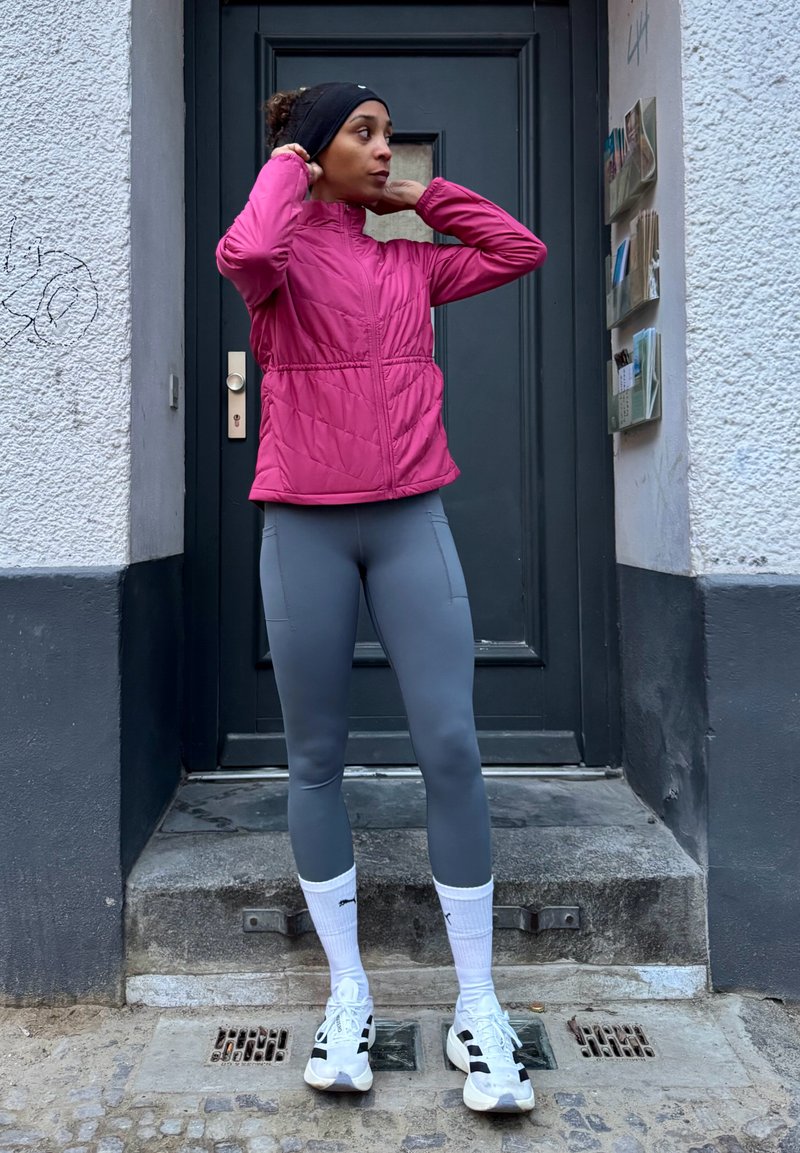 Fitted, insulated pink jacket with a high collar, paired with light gray leggings. White athletic socks and black and white sneakers. Outdoor setting.