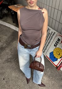 Rjav prilegajoč top z ramenskimi detajli, modri široki kavbojki, rjav usnjen pas in majhna, teksturirana torbica. Siva asfaltna podlaga.