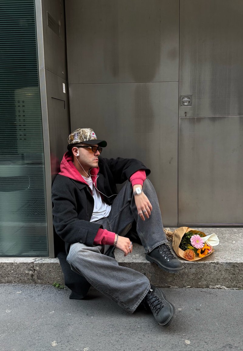 Young man wearing a cap and headphones, sitting on a sidewalk ledge next to a bouquet of flowers, leaning against a metal wall.