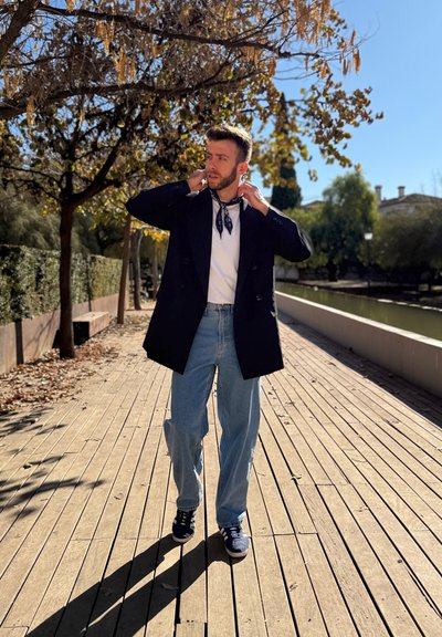 Abrigo negro de doble botonadura sobre una camisa blanca, pañuelo azul al cuello, jeans claros holgados y zapatillas navy, de pie sobre un camino de madera.