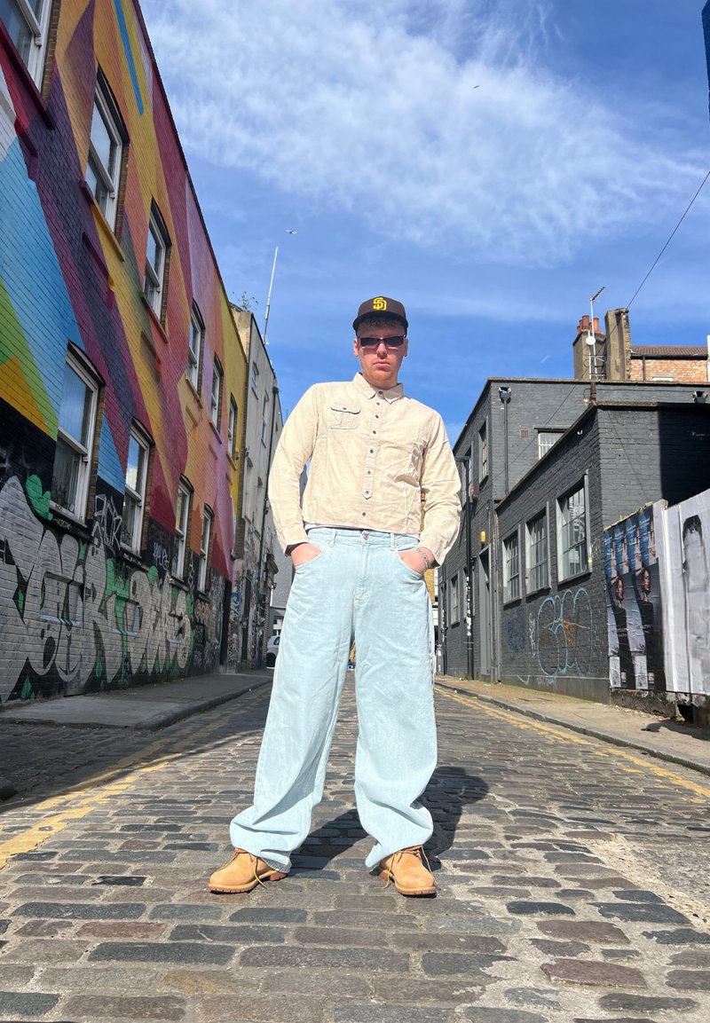 Μπεζ πουκάμισο με κουμπιά, ανοιχτό μπλε φαρδιά τζιν και καφέ παπούτσια. Πίσω έχει πολύχρωμα γκράφιτι σε τοίχους από τούβλα.