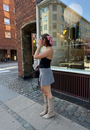 Una mujer está de pie en una calle de la ciudad, vistiendo una camiseta negra sin mangas, una minifalda gris y botas de rodilla color beige, sosteniendo un pequeño bolso.