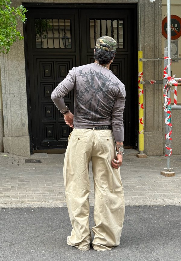 Hombre que lleva una gorra de camuflaje, camisa gris de manga larga y pantalones anchos beige, de pie frente a una puerta negra en una calle de la ciudad.
