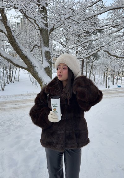Mujer con abrigo oscuro de piel y sombrero blanco sosteniendo una taza, de pie junto a árboles y una carretera cubiertos de nieve en un escenario invernal.