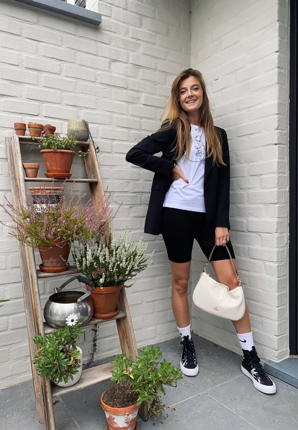 Junge Frau in einem schwarzen Blazer und Shorts, die eine weiße Handtasche hält, steht neben einer Holzleiter mit mehreren Topfpflanzen und einer Gießkanne.