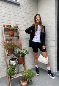 Junge Frau in einem schwarzen Blazer und Shorts, die eine weiße Handtasche hält, steht neben einer Holzleiter mit mehreren Topfpflanzen und einer Gießkanne.