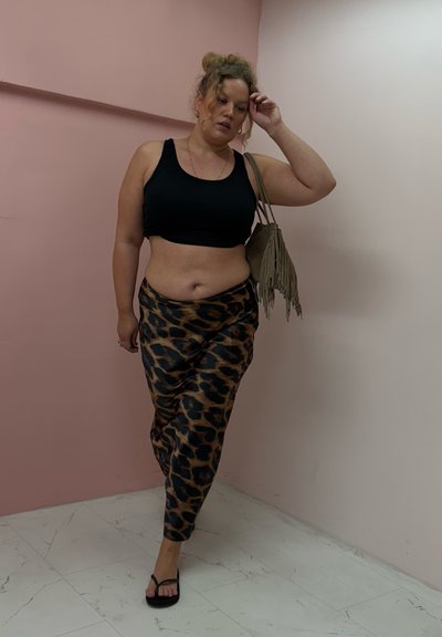 Mujer con cabello rizado que lleva una camiseta de tirantes negra, pantalones de leopardo, chanclas y que lleva un bolso con flecos, caminando en interiores contra paredes rosas.