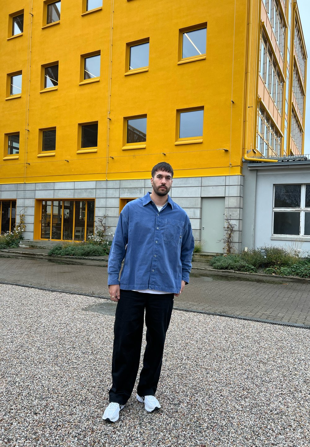 Άνδρας με γένια και κοντά μαλλιά, ντυμένος με μπλε πουκάμισο, μαύρο παντελόνι και λευκά παπούτσια, στέκεται σε χαλίκι μπροστά από κίτρινο κτίριο.