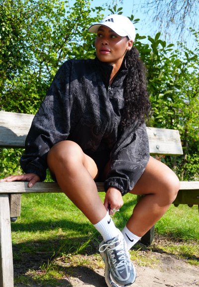 Mujer con una gorra blanca, chaqueta negra y zapatillas sentada en un banco de madera al aire libre ajustándose la media, rodeada de follaje verde.