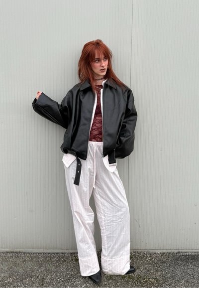 Joven con cabello rojo que lleva una chaqueta negra oversized, top de encaje burdeos, pantalones anchos blancos y zapatos negros, de pie contra una pared gris.