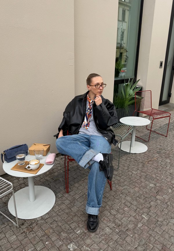 Personne portant des lunettes, une veste en cuir, un jean et une écharpe assise, les jambes croisées, à une table de café en plein air avec un café et un verre d'eau.