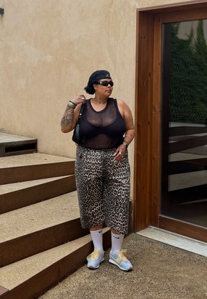Top sin mangas de malla negra, pantalones de pierna ancha con estampado de leopardo, zapatillas deportivas azules con detalles amarillos, boina negra y gafas de sol; de pie junto a una puerta de madera.