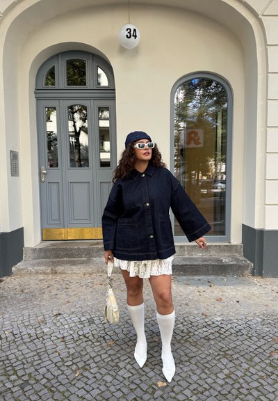 Chaqueta de mezclilla oscura de gran tamaño con botones grandes, combinada con un vestido mini de encaje blanco, calcetas blancas hasta la rodilla y zapatos blancos de punta.