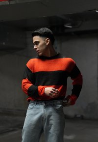 Μαύρο και κόκκινο ριγέ πλεχτό πουλόβερ με μακριά μανίκια, συνδυασμένο με ανοιχτό μπλε τζιν. Η υφή φαίνεται απαλή και άνετη.