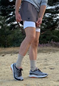 Pantaloni sportivi grigi sovrapposti a pantaloni compressivi bianchi, abbinati a scarpe sportive grigie e calze grigie chiare, ambientati in un'area esterna sabbiosa.