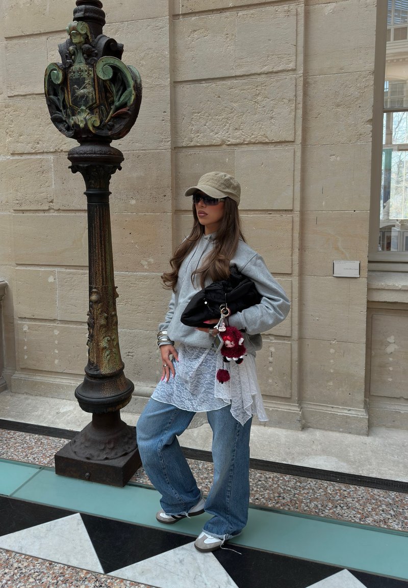 Donna con berretto, occhiali da sole, felpa grigia, top in pizzo, jeans larghi e sneakers, in piedi all’interno vicino a un palo di metallo ornato su pavimento piastrellato.