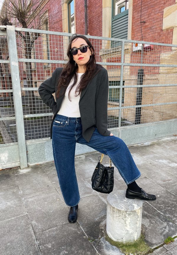 Blazer preto sobre uma camisa branca, combinado com jeans azuis de perna larga. Os acessórios incluem óculos de sol pretos e uma mala preta acolchoada.