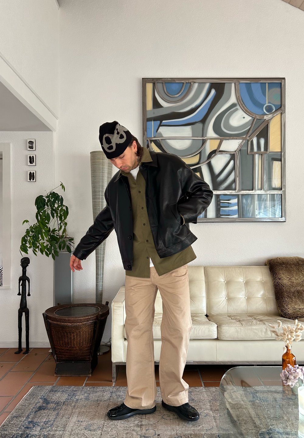 Veste en cuir noire sur une chemise vert olive, pantalon beige et chaussures noires. Le décor comprend un canapé blanc et une œuvre d'art murale abstraite.
