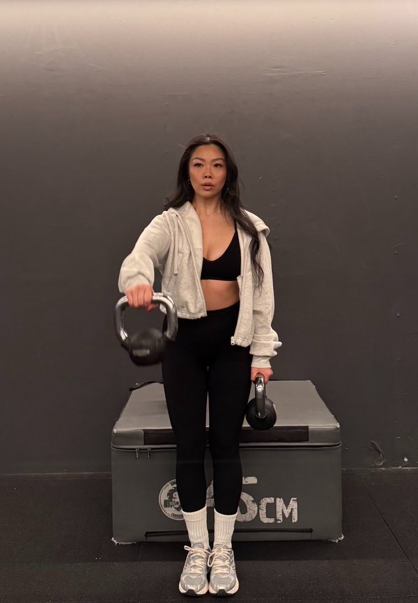 Svart kettlebell holdt i høyre hånd, grå hettejakke med glidelås, svart sports-BH og leggings. Grå plattform med merke i bakgrunnen.