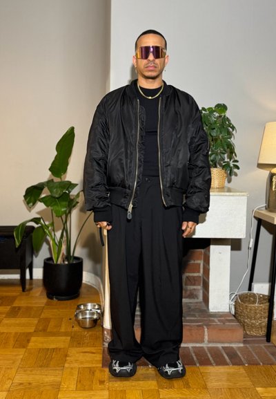 Hombre con chaqueta y pantalones negros de gran tamaño, gafas de sol futuristas con visera tintada, de pie en interiores cerca de plantas y una lámpara.