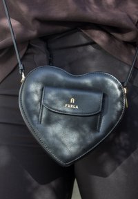 Čierna kožená crossbody taška vo tvare srdca značky Furla, nosená na úrovni pásu cez tmavé oblečenie.