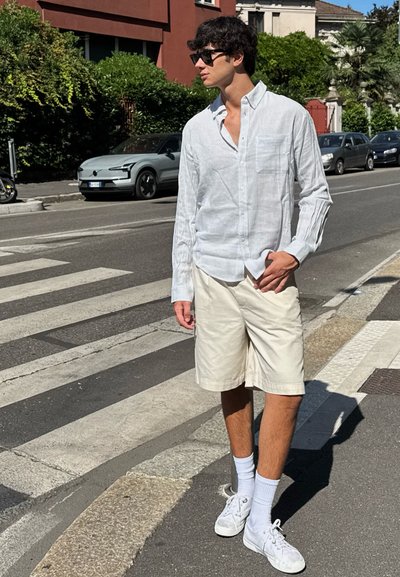 Camisa de rayas blancas de manga larga con un bolsillo, pantalones cortos beige, calcetines blancos y zapatillas, de pie en un paso de peatones en un entorno urbano.