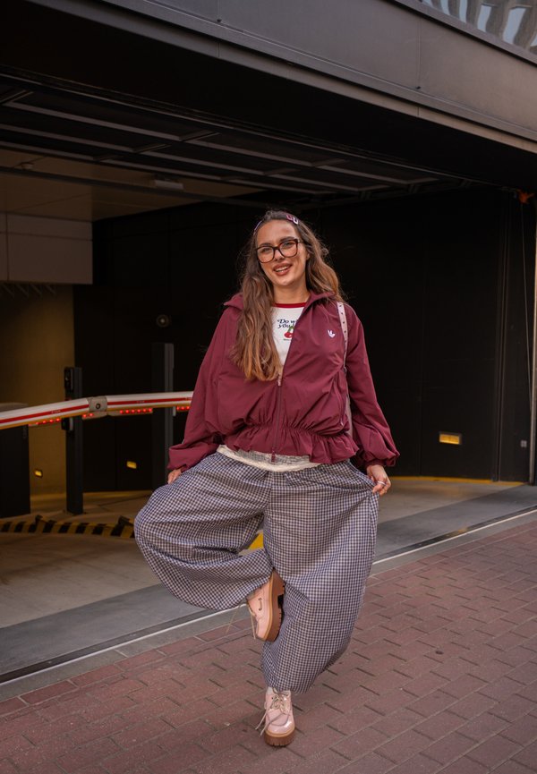 Una mujer sonriente con gafas, una chaqueta color burdeos y pantalones anchos de cuadros posa delante de un edificio, levantando una pierna para mostrar un zapato rosa.