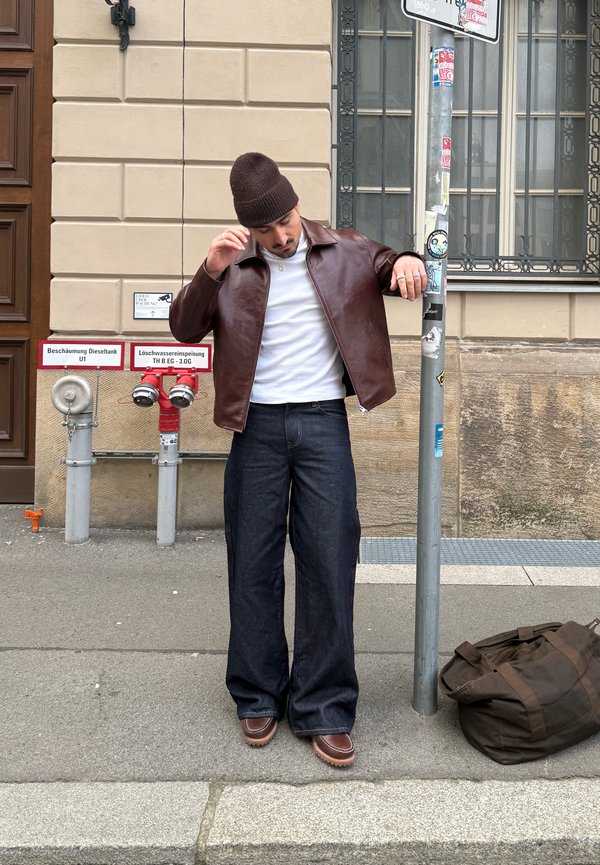 Uomo con berretto marrone e giacca di pelle che sistema il colletto, in piedi sul marciapiede accanto a un palo e una borsa marrone vicino al muro di un edificio.