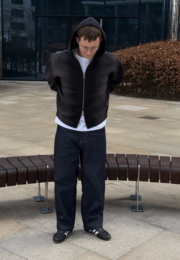 Jonge man in een zwarte hoodie, wit shirt, donkere spijkerbroek en zwarte schoenen staat naar beneden kijkend in een stedelijk plein met een gebogen houten bank achter hem.