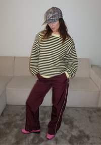 Pull rayé jaune et gris foncé, pantalon de survêtement bordeaux avec bandes latérales, chaussures roses et casquette camouflage, debout sur un tapis gris.