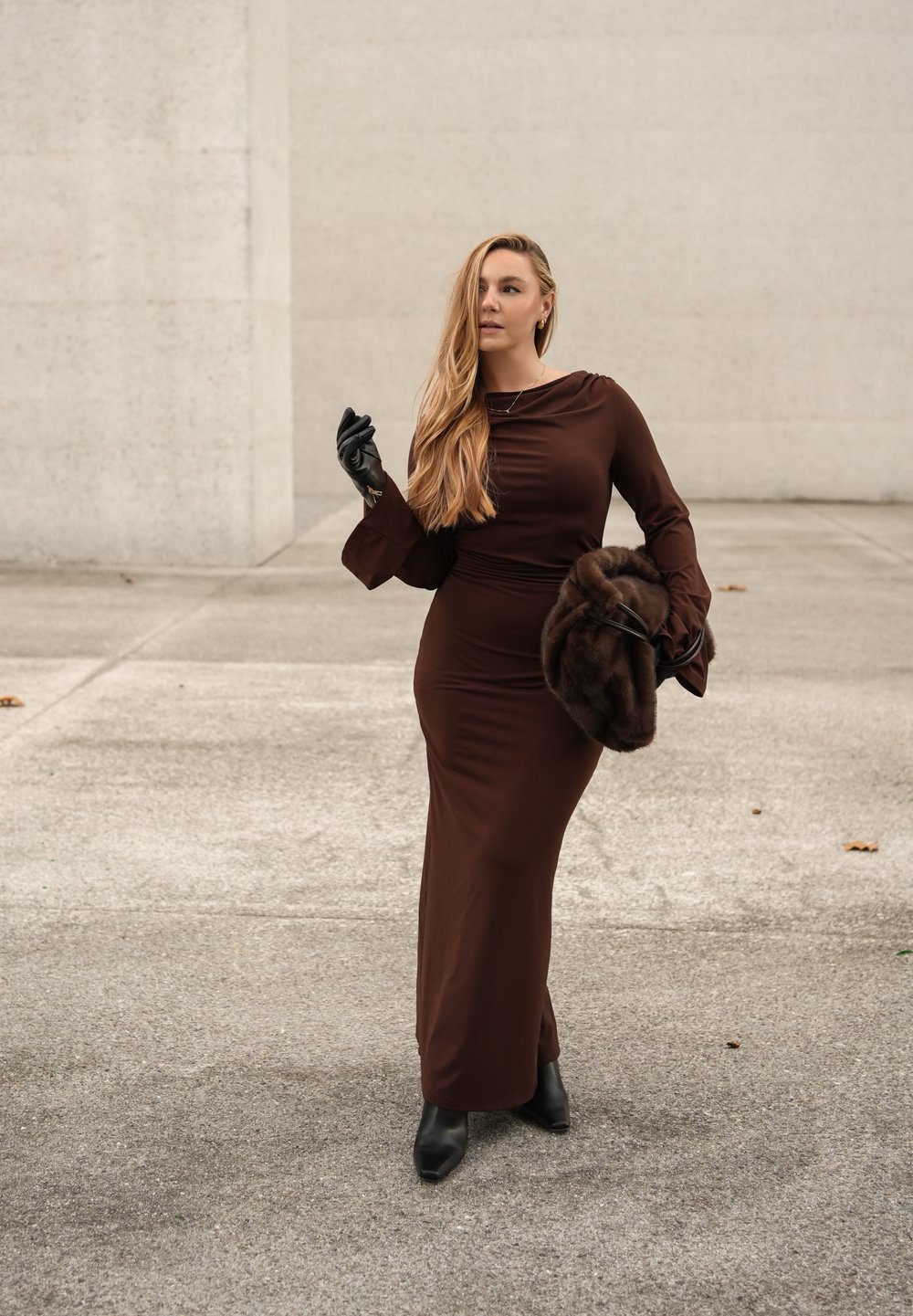 Brun kjole med lange ærmer, udvidede manchetter, figursyet design og rynket halsudskæring. Holder en mørk pelsstola, iført sorte handsker og støvler.