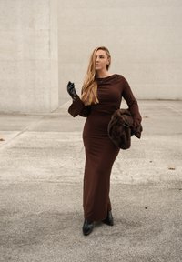 Brun kjole med lange ærmer, udvidede manchetter, figursyet design og rynket halsudskæring. Holder en mørk pelsstola, iført sorte handsker og støvler.