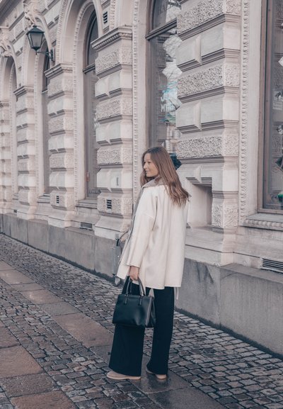 Abrigo blanco de gran tamaño, pantalones negros de pierna ancha y bolso negro. Pavimento de adoquines y edificio ornamentado en el fondo.
