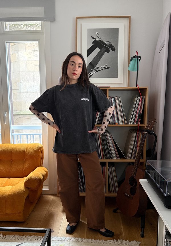 Femme debout, mains sur les hanches, dans un salon, portant une chemise noire ample avec des manches en tulle à pois, un pantalon marron, près d'un fauteuil jaune et d'une guitare.