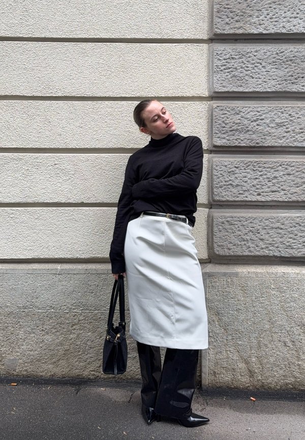 Kvinde iført sort rullekrave, hvid nederdel over sorte bukser, holder en sort taske, lænet op ad en struktureret grå og beige stenmur.