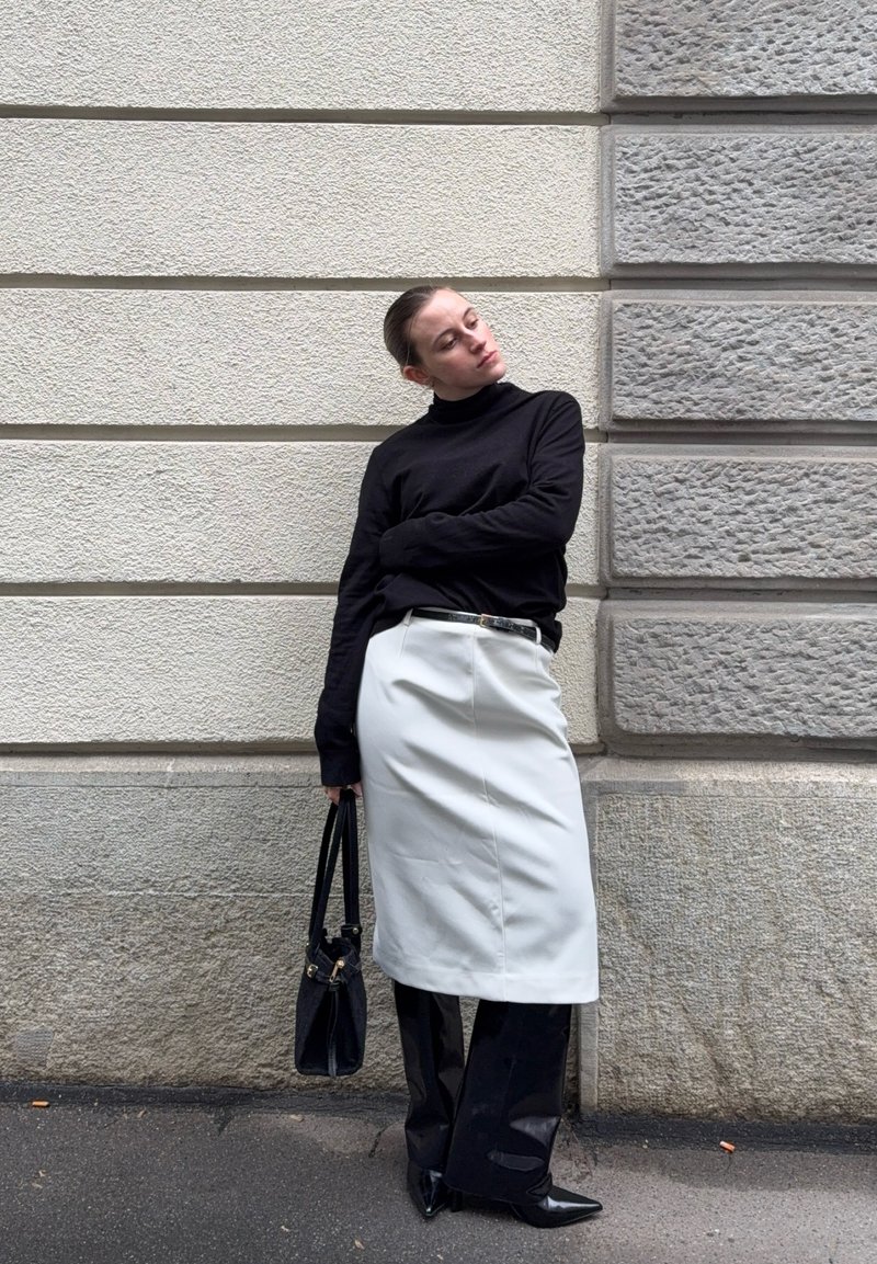 Γυναίκα με μαύρο ζιβάγκο, άσπρη φούστα πάνω από μαύρο παντελόνι, κρατώντας μαύρη τσάντα, στηριζόμενη σε υφή γκρι και μπεζ πέτρινο τοίχο.