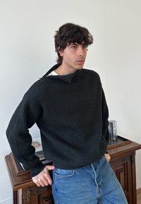 Ung mann med krøllete mørkt hår iført en svart ribbestrikket genser og blå jeans, lent mot et tregskap med en metallkopp på.