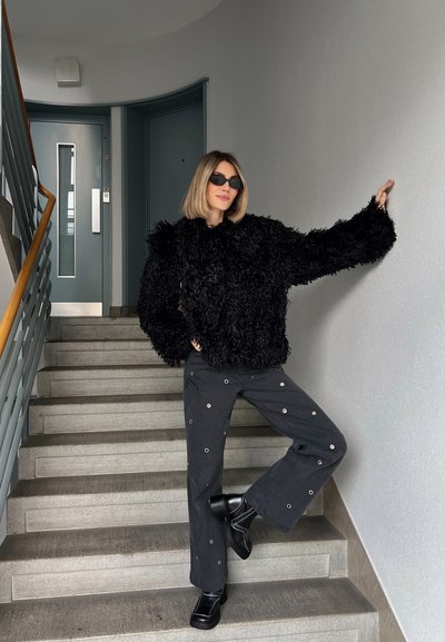 Suéter negro esponjoso, de corte holgado; pantalones grises de pierna ancha con detalles de herrajes circulares; zapatos de plataforma negros; de pie en escaleras beige.