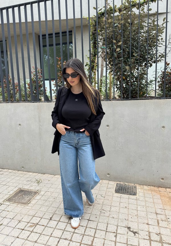 Frau trägt schwarzen Blazer, schwarzes Hemd, weite blaue Jeans, weiße Sneakers und Sonnenbrille, geht auf einem gefliesten Gehweg an einem Metallzaun und Sträuchern entlang.