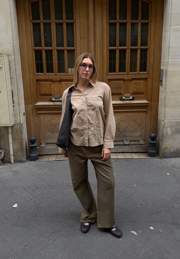 Une personne portant des lunettes, une chemise à carreaux et un pantalon ample se tient sur un trottoir en ville, devant des portes doubles en bois, avec un sac noir en bandoulière.