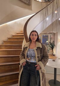 Jeune femme portant un manteau en peau retournée et un haut à motifs, debout près d'un escalier en bois courbe à l'intérieur d'un bâtiment moderne et doucement éclairé.