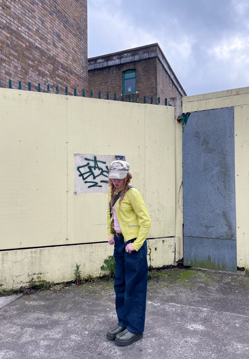 Personne en veste jaune, chemise rose, pantalon large bleu et casquette beige se tient près d’un mur jaune avec des graffitis et des bâtiments en brique en arrière-plan.